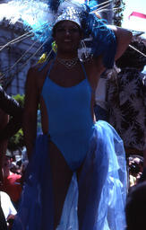 A man in a light blue leotard and large feathered headdress standing on the street.