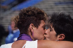 Two women kissing and embracing, one has a purple ribbon around her neck.