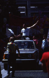 Supervisor Harvey Milk sitting on the roof of a car driven by campaign manager, Anne Kronenberg, through the June 25, 1978 Gay Freedom Day parade with a sign that says "I'm from Woodmere, N.Y." and waving to the crowd. 