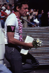 Supervisor Harvey Milk leaning up against a vehicle waiting to speak at Civic Center Plaza on Gay Freedom Day, June 25, 1978. Supervisor milk wears a white ringer t-shirt with red trim, a pink lei, a black armband with a pink triangle, and holds a bunch of daises in one hand.