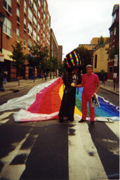 A snapshot of Gilbert Baker with a costumed companion and a very large rainbow flag, apparently one of the multi-block flags Baker created for Pride parades.