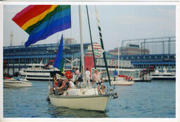 A snapshot of Gilbert Baker and companions on a sailboat which is flying a large rainbow flag.