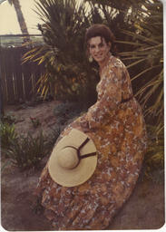 D'Anne at a photoshoot outdoors. She is wearing a long, flowing dress and is holding a straw sunhat.